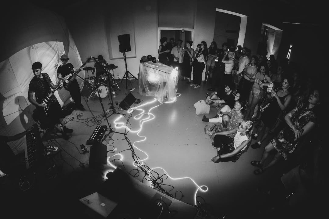 Una banda tocando en un extremo de la sala, el público sentado en el piso alrededor y una instalación de luz cruzando el suelo