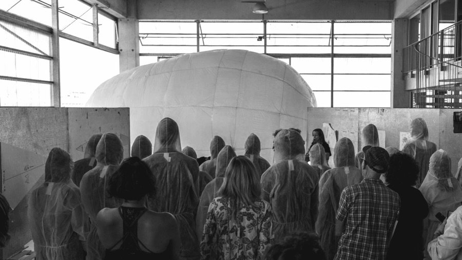 Un grupo de personas con monos blancos de espaldas, mirando una gran cúpula inflable dentro de una nave